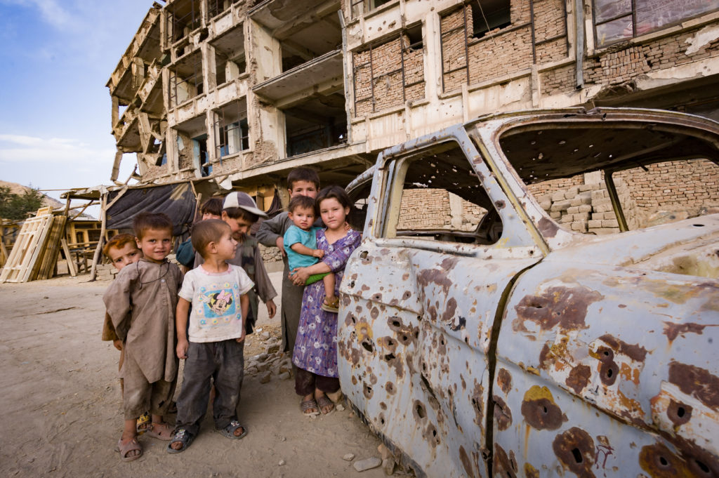 kids in front of shot up rusty car in destroyed town